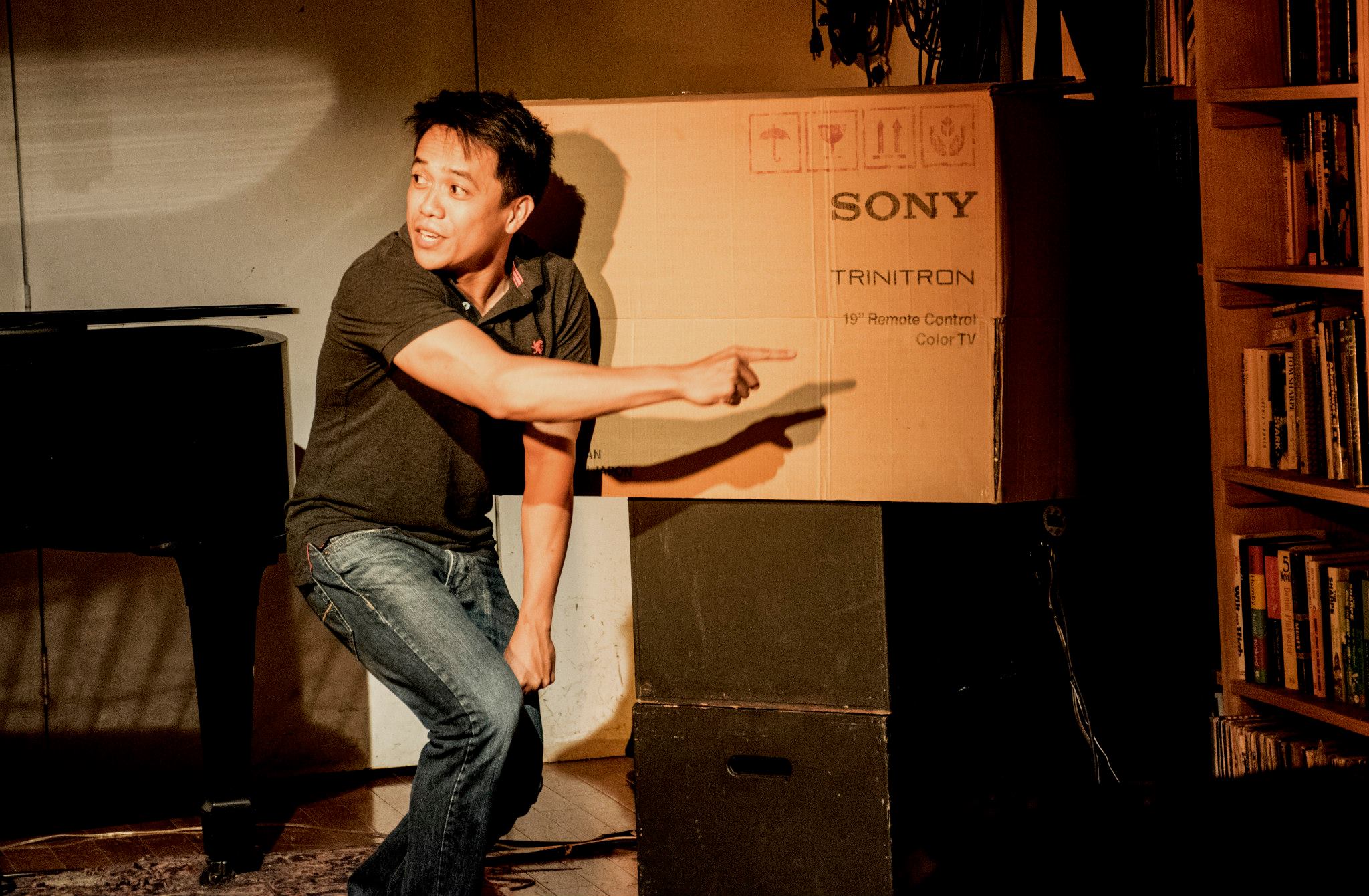 a Filipino man stands in front of a grand piano and a cardboard television box