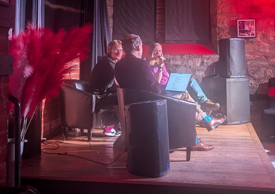 a photo of three people onstage at a live podcast recording. Josh sits in the middle next to his cohosts A.J. and Brian.