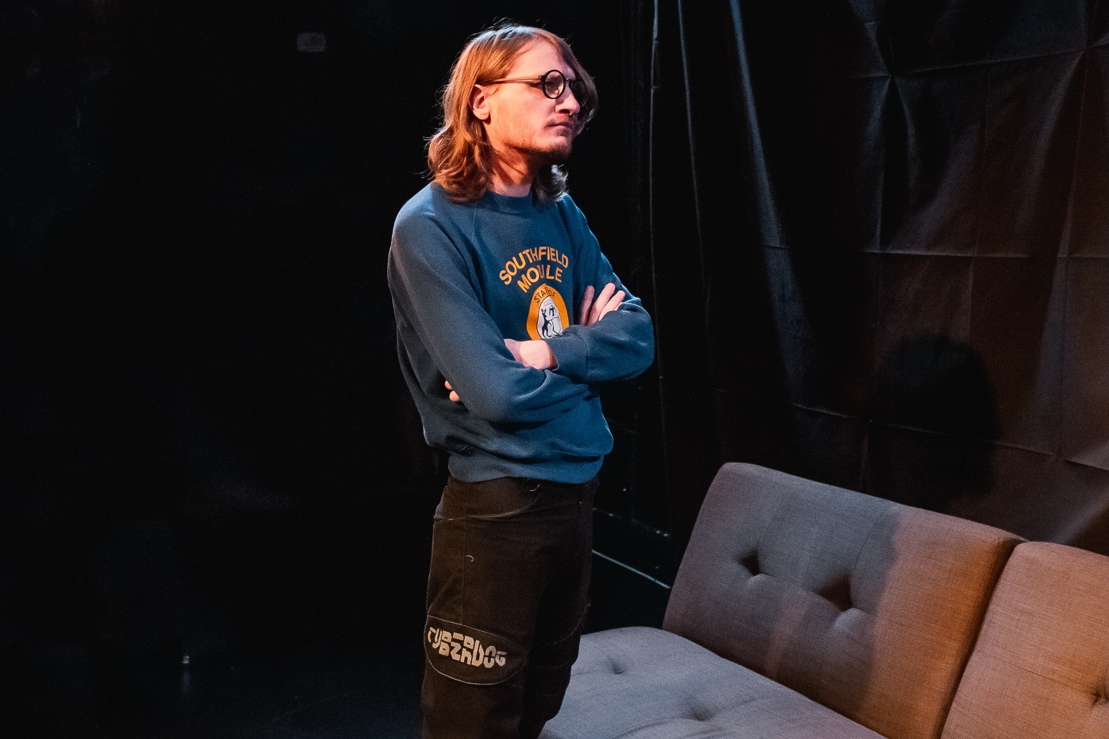 a photo of josh, a blond wearing glasses, a blue sweatshirt, and black raver pants. he is looking off into the middle distance