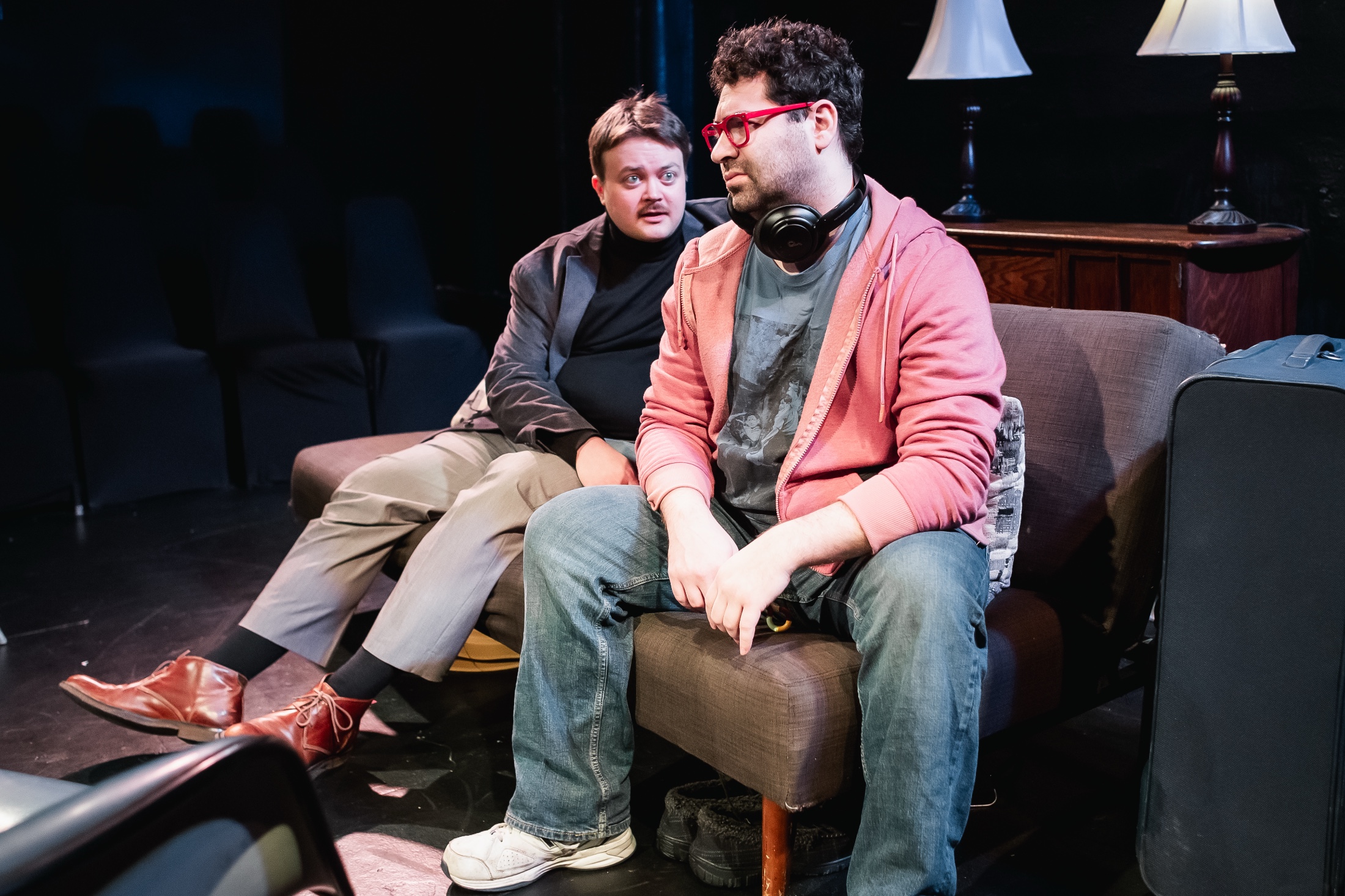 a photo of two actors in a play. the actor closer to the camera wears glasses and has curly brown hair, is wearing a hoodie, jeans, and a t-shirt, and is looking off in disdain.
the actor behind them has brown/blond hair, is wearing casual business attire, and seems to be trying to explain something